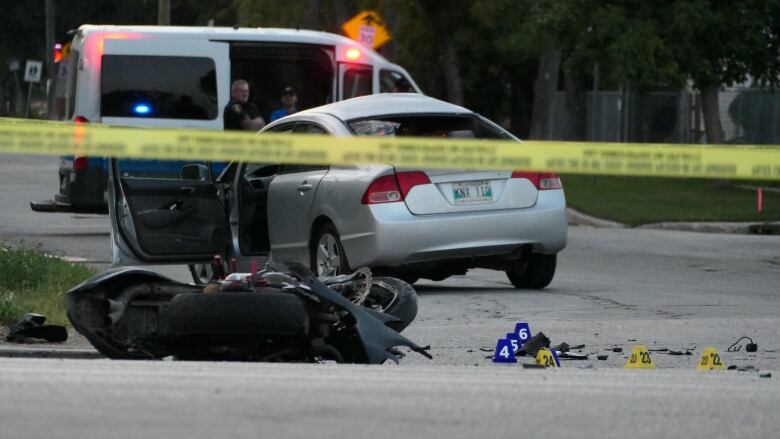 A crash motorcycle lies on its side on a street. A car with an open door sits in the background. Yellow police tape blocks off the scene.