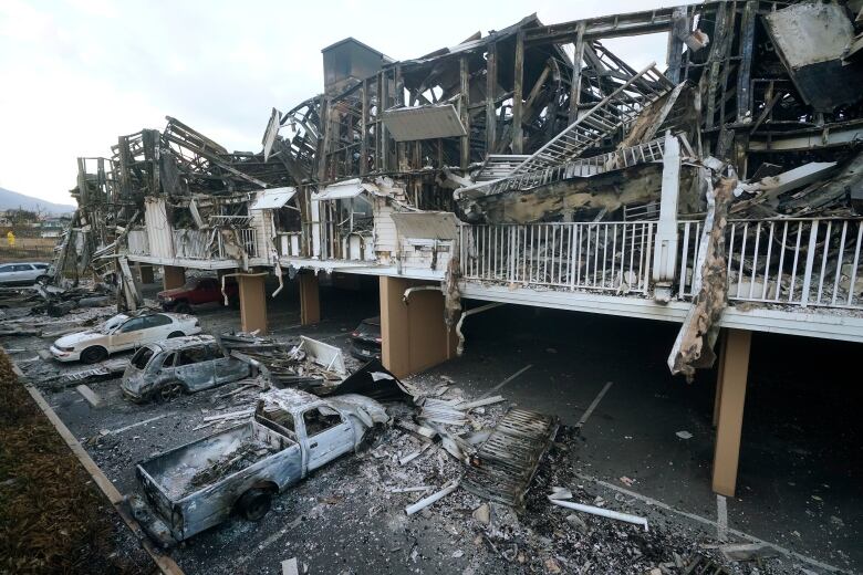 Burned out vehicles with no tires sit in front of burned, twisted structure.