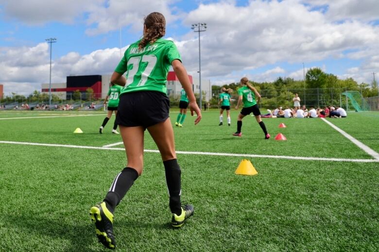 Girls are wearing bright green and white soccer jersey's. They are warming up for their game on a turf field with cones set in place for drills. 