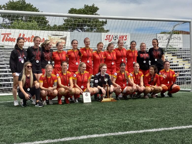 Women in red soccer uniforms in front of a net with trophy.