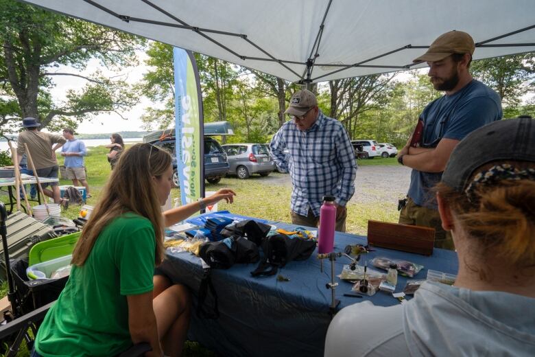 A woman sits behind a table while two men loot at objects on the table.