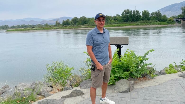 A man in grey polo shirt and shorts stands in front of a river.