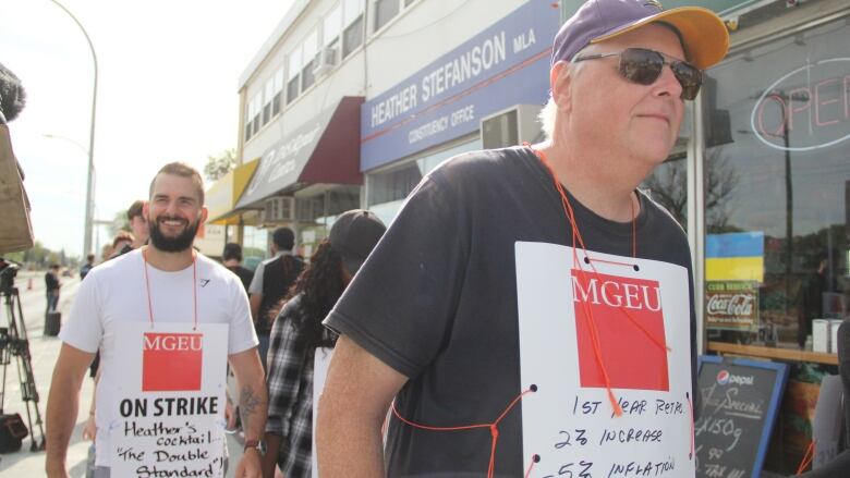 Two people walking with picket signs hanging on the front of their bodies.