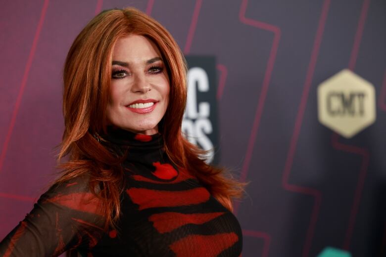 Shania Twain, with long red hair and a black and red dress, stands in front of a media backdrop, looking at the camera.