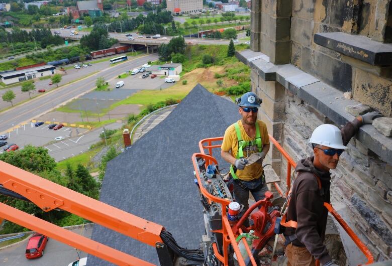 Two construction workers in a bucket truck full of rocks work on the side of an old church 80 feet above the city. 