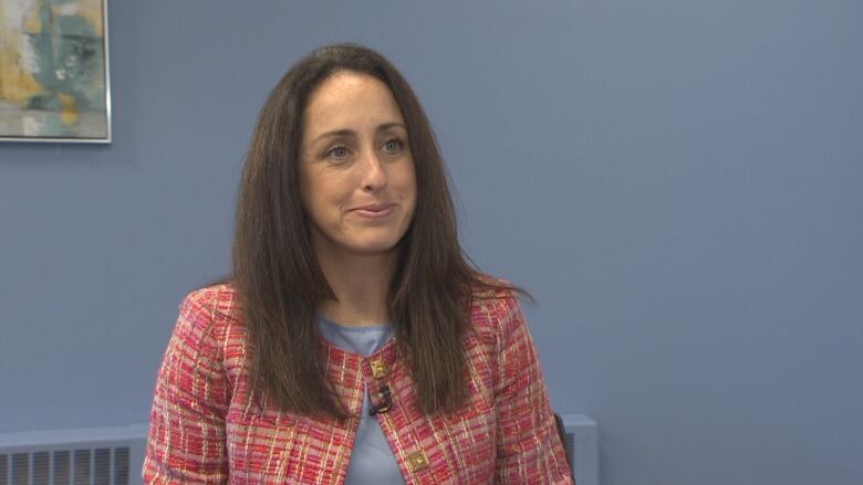 A woman wears a pink tweed jacket with a blue shirt sits in front of a blue wall.