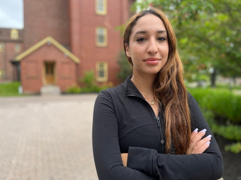 A young woman wears a black zip up sweater with a serious expression against the backdrop of brick buildings. 