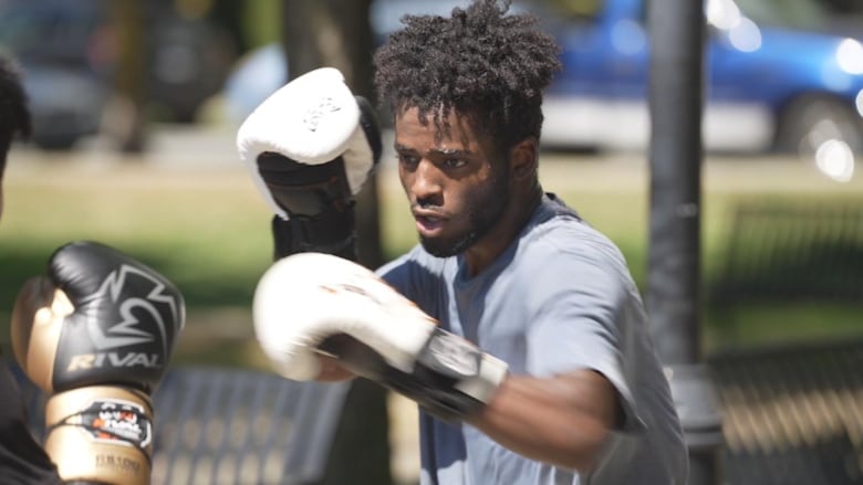 A closeup of a man in dreadlocks sparring with white boxing gloves against an opponent, with a park blurred out in the background.