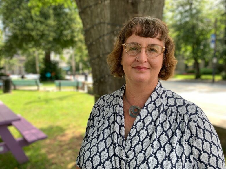 A non-binary person wearing glasses stands in a shady park by a downtown street.