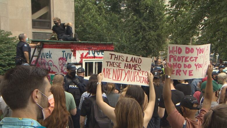 A police negotiator speaks with a protester on the roof of a wooden shelter during a protest in Halifax in Aug. 2021. People hold up signs in the foreground of the photo protesting the removal of the structure. 