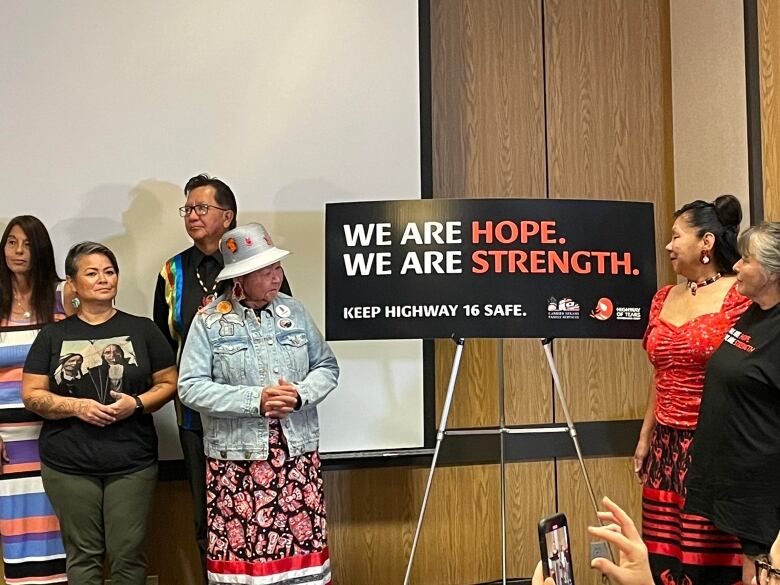 Indigenous people, some wearing ribbon skirts and traditional jewelry, stand by a sign that's just been unveiled. The black, white, and red sign says, 'We are hope. We are strength. Keep Highway 16 safe.'