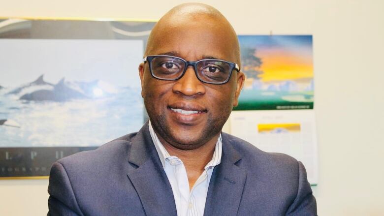 A Black man wears glasses and a navy blue suit.