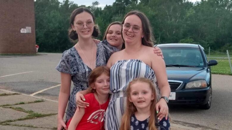 A smiling woman wearing glasses and summer clothes is surrounded by three girls and a tall young woman on a sidewalk.