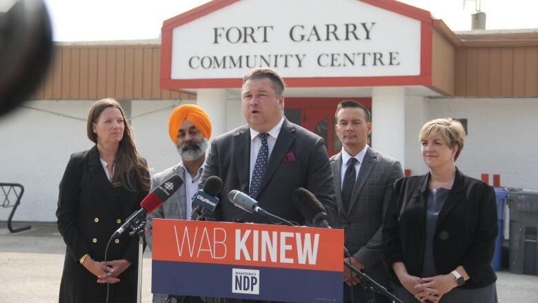 Several politicians stand behind a podium, in front of the front entrance of a community centre.