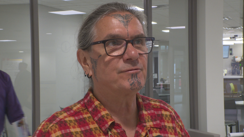 Man pictured from shoulders up wearing a red and yellow patterned shirt, glasses and cultural markings on his face.