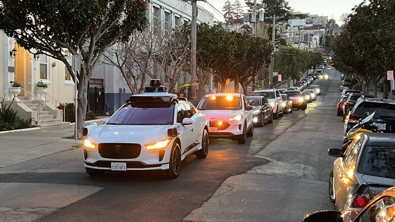 A long line of cars on a San Francisco street, with a white driverless taxi at the front.