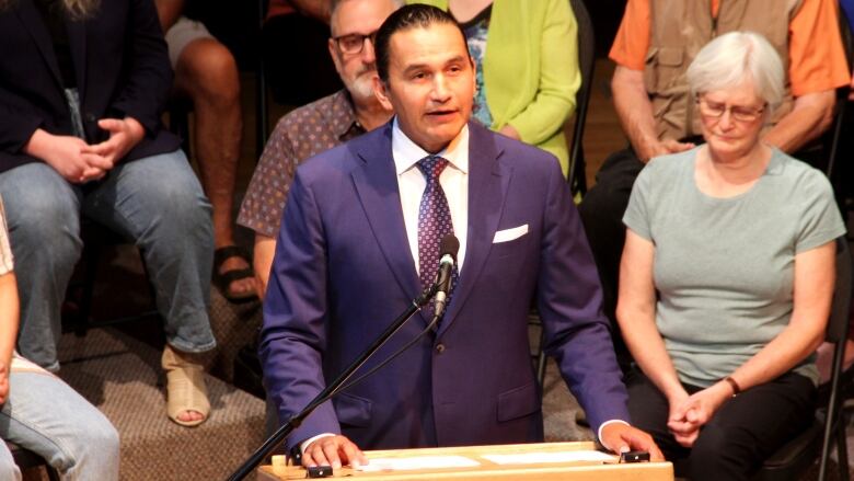 A man in a suit speaks behind a podium while several people sit behind him. 