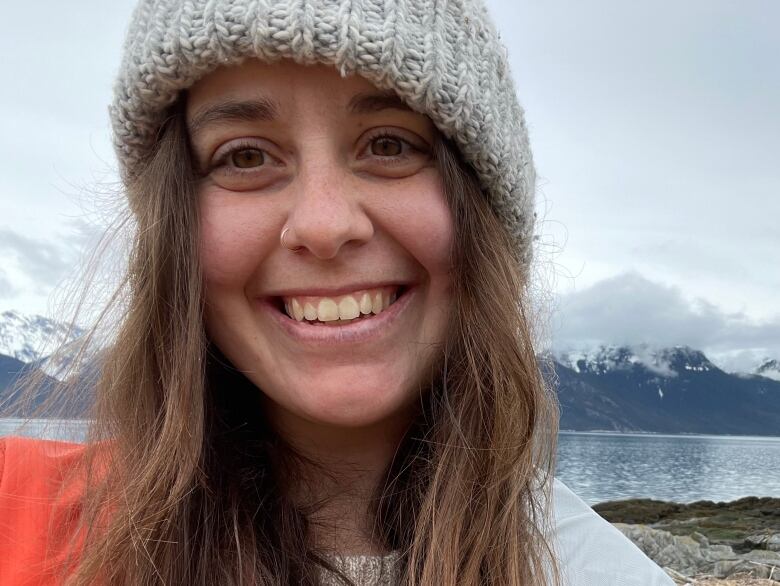 A selfie of a smiling in a toque standing by the water with mountains in the background.