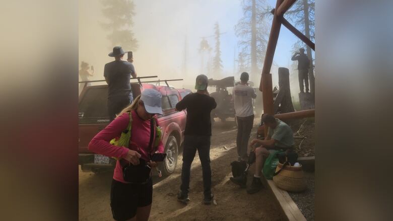 People stand on the road with vehicles behind them and trees on both sides of the road.