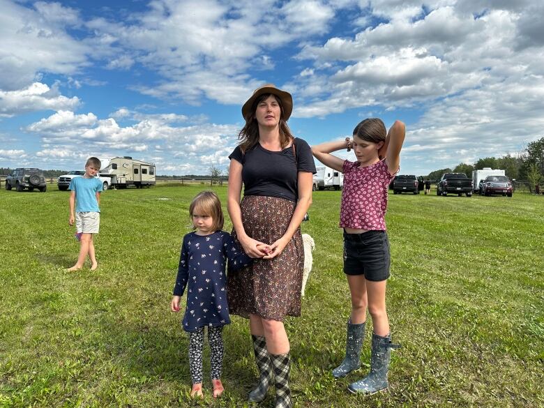 A woman in a dress and a hat stands with two young girls outdoors, with a boy behind her in the background.