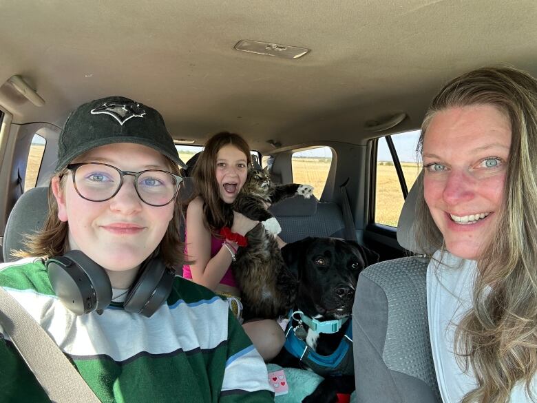 A family, three women, a dog and a cat, sit in a vehicle with their packed belongings.