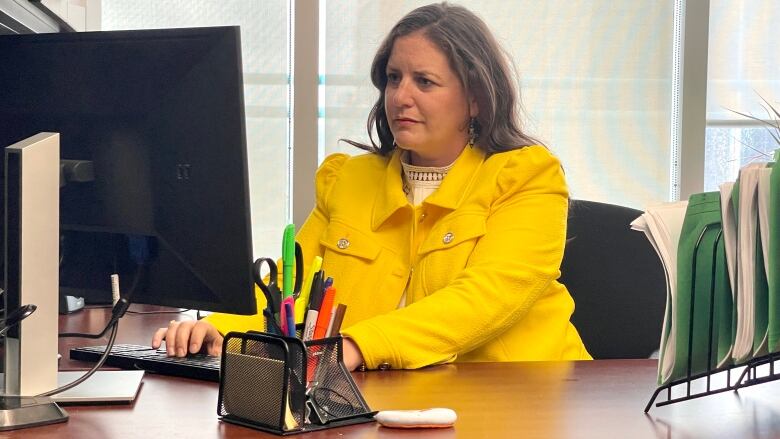 A Caucasian woman in a yellow blazer sits at a desk working on a computer.