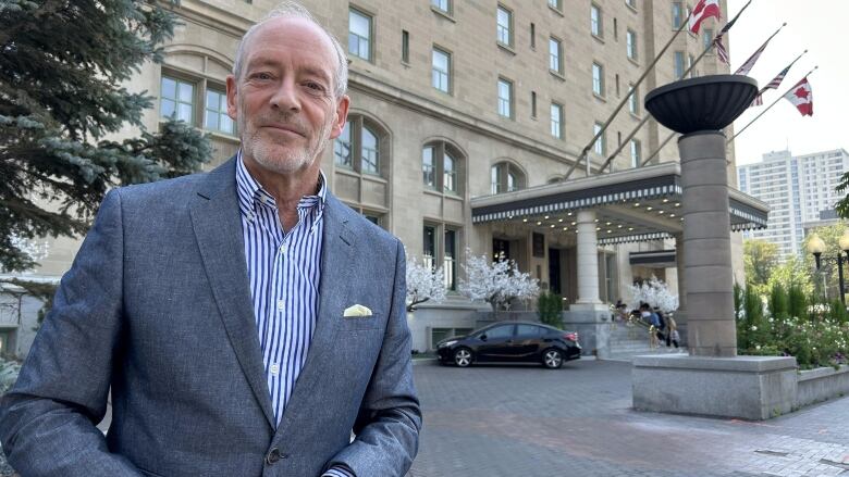 A man wearing a suit looks to the camera in front of a hotel.