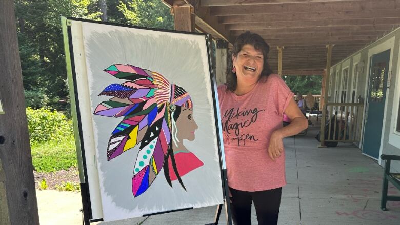 A woman wearing a peach-colour t-shirt stands next to a colourful painting of an Indigenous woman wearing a feathered headdress. 