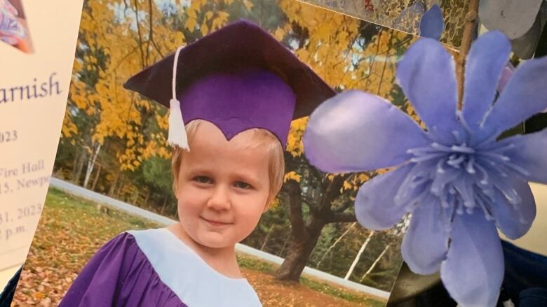 A young boy wearing a purple graduation cap and gown is seen in a photograph. A purple flower rests next to the photo.