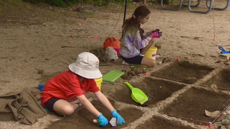 Two young girls are digging in the dirt searching for artifacts. One is wearing a red shirt, white hat and blue gloves. The other is wearing a purple sweater and pink gloves. 