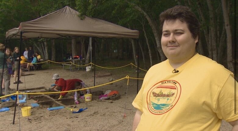 A man with brown hair smiles. He is wearing a yellow t shirt. He stands in front of a tent where kids are digging for archaeological artifacts. 