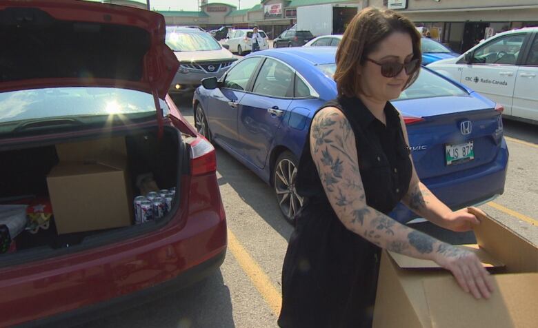 A woman in sunglasses and a black dress stands behind a red car while folding the flaps on a cardboard box