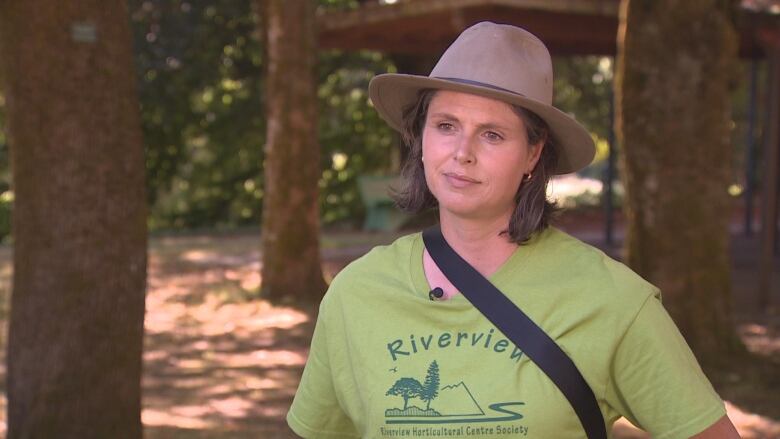 A woman stands surrounded by trees in a green shirt that says 'Riverview Horticulture Centre Society.'