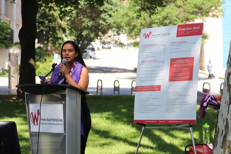 Woman speaking into a microphone in a park with a board of statistics beside her. 