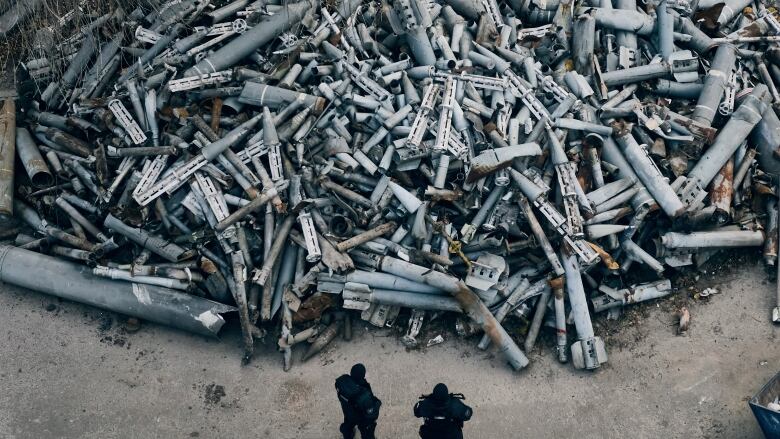 Two people stand in front of a large pile of spent rockets and bombs. 