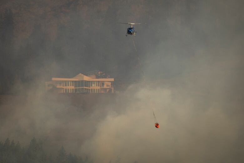 A helicopter flies through smoke-choked skies with a home in the background visible through the haze.