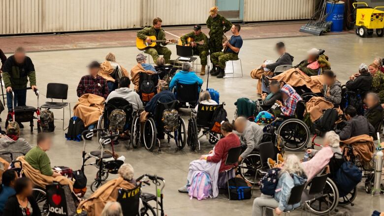 People in camouflage play guitars in a hangar, surrounded by people in wheelchairs.