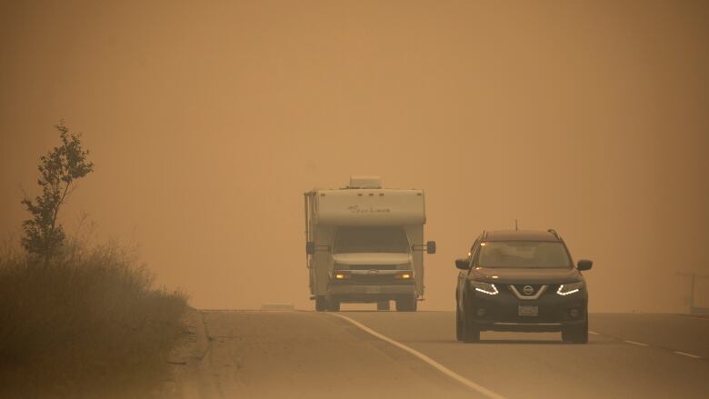 A car and a truck are seen driving away from thick wildfire smoke.