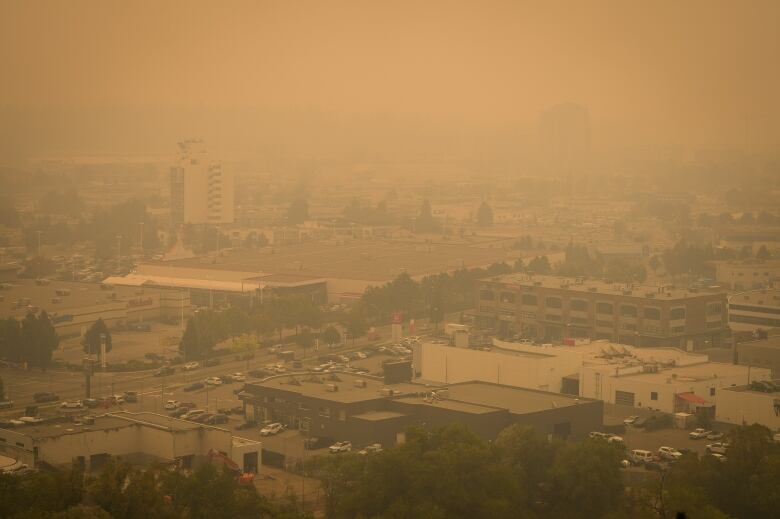 Looking down at low-rise buildings with parking lots, and two high-rises, all against a brown murky sky with smoke obstructing the field of view.