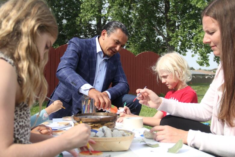 A man pours paint from a bottle onto a table. Some kids and a woman are working on arts and crafts around the table 