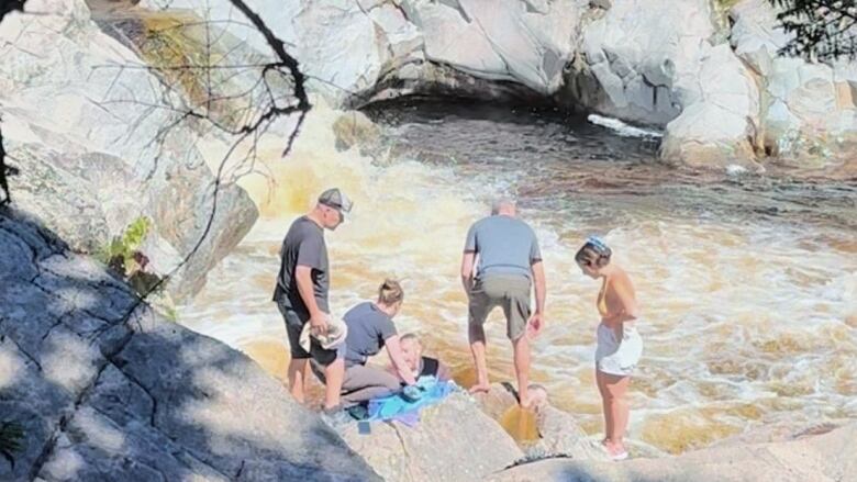 A group of people stand on some rocks next a gushing river. One person can be seen in the water.