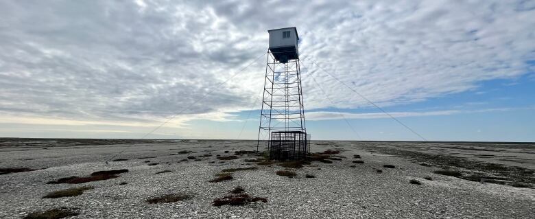 A wooden structure sits atop a tower held by wires on a beach ridge, with a blue sky and clouds in the background.