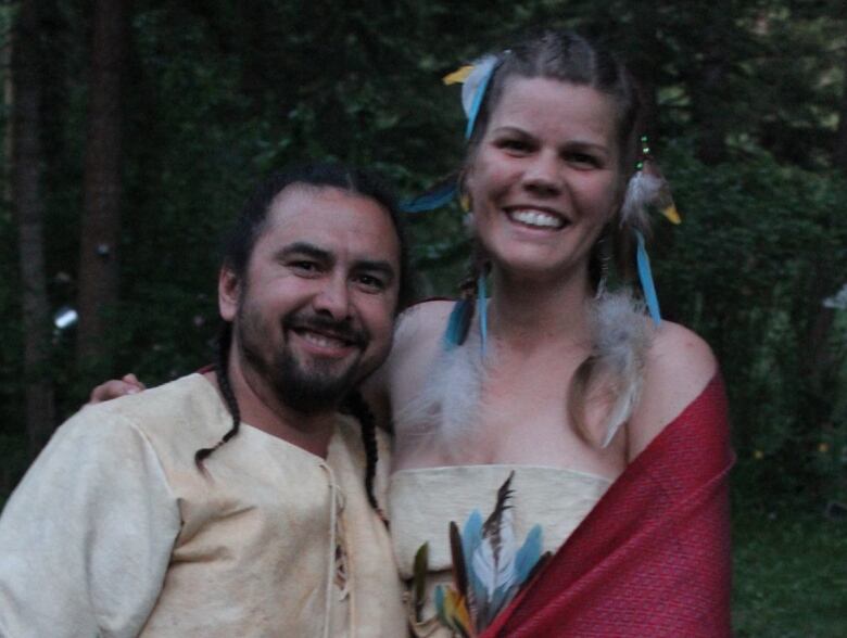 A man and woman stand outside, they are dress in wedding clothes, embracing and smiling at the camera. 