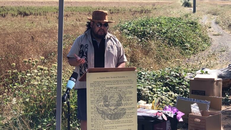 A man in a hat and sunglasses stands behind an outdoor podium delivering a speech.