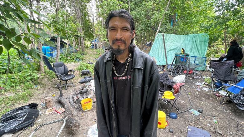 A man with long hair and a leather jacket is looking at the camera. Behind him are tents, garbage and debris on the ground, surrounded by trees.