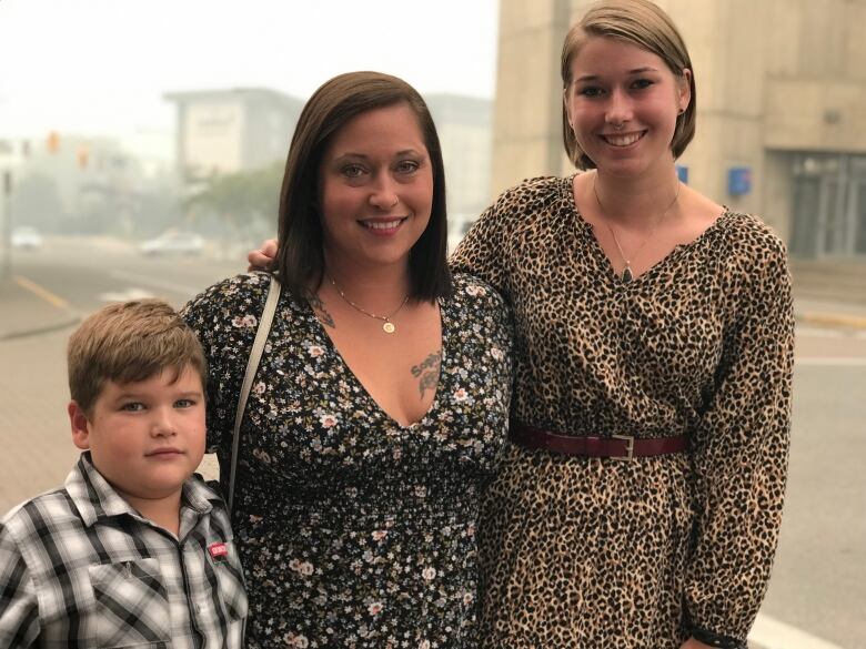 A woman in a floral dress stands for a posed photograph with her teen daughter in a similar outfit and her younger son, who wears a plaid shirt.