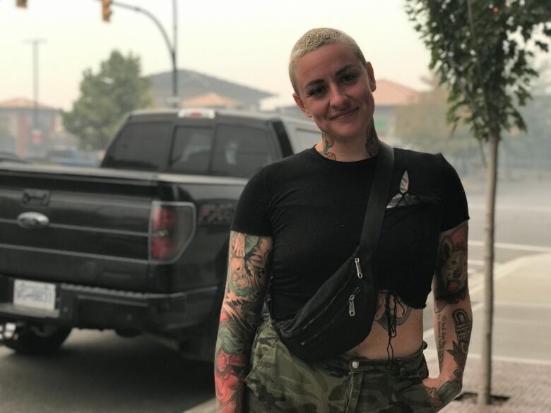 A woman poses for a photograph with short dyed-blonde hair, a black shirt, and tattoos on her arms.