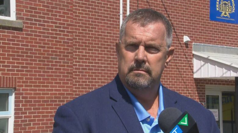 Man with short grey-brown beard and moustache wearing blue suit jacket standing outside brick building.