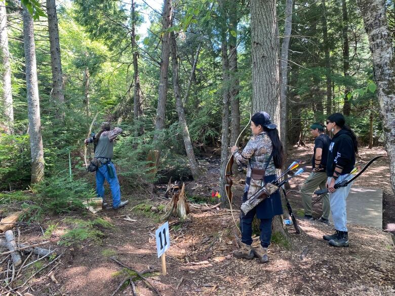 a man using his bow to hit a target with 3 other people watching while in the forest. 