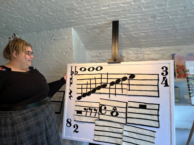 Person in a black shirt and plaid skirt stands beside a large felt board with felt music notes on it. 